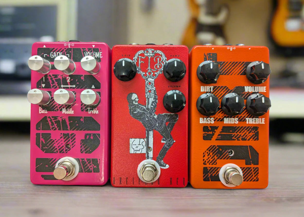 Three Funny Little Boxes guitar effect pedals on a wooden surface with a blurred guitar background.